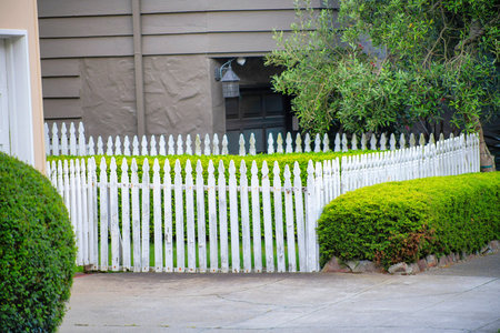 White picket fence in front of house or home in the neighborhood with square and rounded front yard hedges. In the city or in the suburban areas of the town with grass and background house.の写真素材