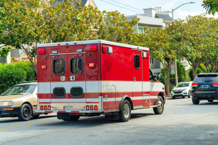 Red ambulance as it speed through the downtown city streets in midday sun with trees and foliage in the background and other cars in foreground. Late afternoon on a sunny day to help an emergency.の写真素材