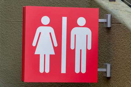 Red bathroom sign with woman and man pattern in white paint on metal post indoors with beige stucco wall in shade. In the city in a building for using restroom with toilet sink and urinal.の写真素材