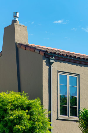 Brown stucco house with visible window and rain gutter pipes with chimney and front yard trees with...