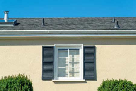 Lone window on beige house facade with green shutters and white accent color with dark roof tiles and vents. Front yard shrubs on suburban home with blue sky background in downtown neighborhood.の写真素材