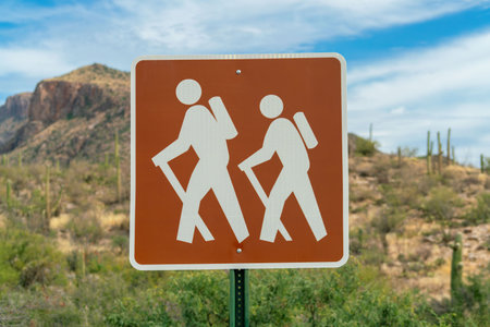 Hiking trail sign with two white stick figures on walk with brown pain square shape for park or outdoor travel. Recreational area in the hills of arizona in late afternoon sun with white clouds.の写真素材