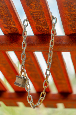 Pad lock with metal chain links tied together and hanging from bars or pieces of wood on gazebo or outdoor roof lining. Visible blue sky and vegetation with blur in afternoon shade.の写真素材