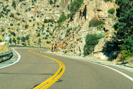 Hot summer road in the hills and mountains of arizona for vehicle and car transportation with metal gaurd rail and rock faces. Afternoon sun with asphalt and yellow and white paint lines.の写真素材