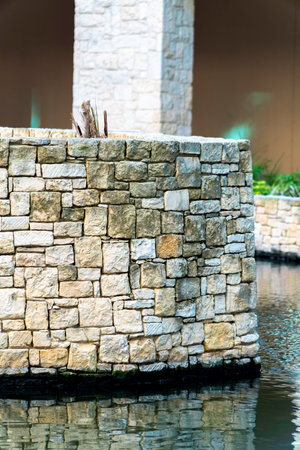 Stone wall with beige white decorative bricks and rocks in afternoon shade near pool or canal in...