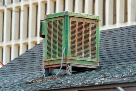 Swamp cooler air condition system on rooftop in modern city with green color and aged and weathered metal body. Gray roof tiles with white cement building background in afternoon shade.の写真素材