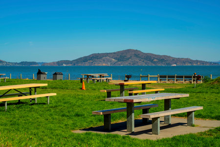 Outdoor park area with several wooden picnic benches and tables for eating and recreation in field with backgrund mountains. In afternoon sun with hazy blue copy space sky for sports and activity.の写真素材