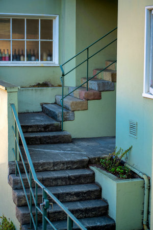 Small entryway to apartment or house in urban area with thin staircase and metal handrail with beige stucco exterior. Late afternoon shade with steps and visible window with decorative bottles.の写真素材