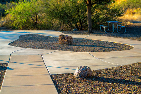 Park or outdoor area with cement sidewalk loop with gravel and stones with background shrubs in sunset light. Trees with decorative landscaping in city neighborhood or suburban park outdoors.の写真素材