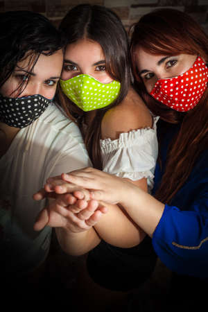Three women friends with colorful face mask protect themselves from coronavirus covid-19 with their hands togetherの写真素材