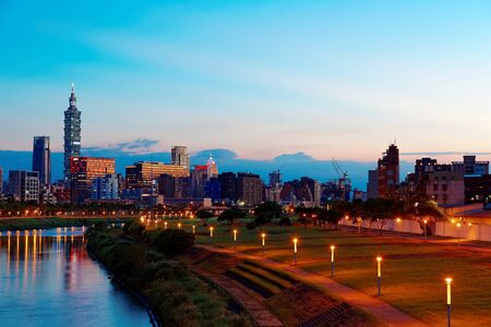 Riverside scenery of Taipei City at dusk with the famous landmark tower amid skyscrapers in Xinyi District Downtown, street lamp lights by the riverbank & reflections on the water of Keelung Riverの写真素材