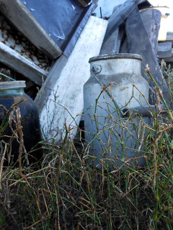 Grunge dirty retro gray milk can with a wooden handle, stands in the grass. Old shabby boards in the backgroundの写真素材