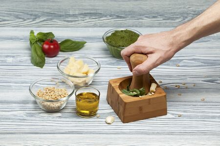 Male hand makes italian pesto in a mortar. Ingredients- basil, parmesan, pine nuts, garlic, olive oil and salt on a light wooden background. Side view. Selective focus. Copy space.の写真素材