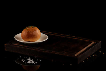 Round wheat bread or bun on a wooden kitchen board. Dark background with copy space. Black glass table with reflection.の写真素材
