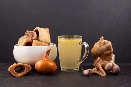 Homemade boiled beef bone broth in transparent cup on a dark background with meat and vegetables....
