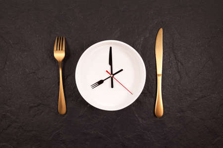 Clock hands on a white plate. Gold fork and knife on a dark stone background. Concept of autophagy or interval fasting.の写真素材