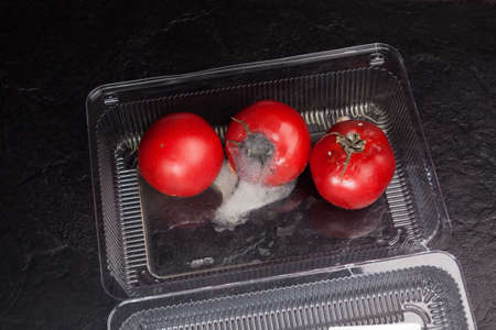 Spoiled tomatoes with mold in disposable plastic container. Organic food waste in supermarkets. Incorrect long-term storage. Dark textured stone background.の写真素材