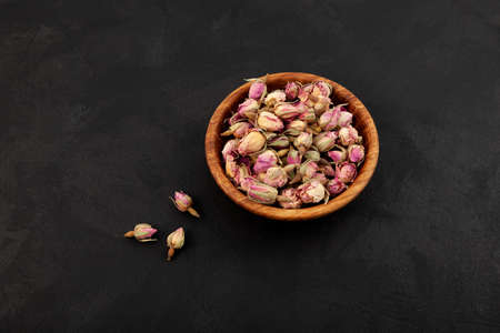 Wooden bowl with Dried pink rose buds on dark textured background. Ingredient for healthy herbal...