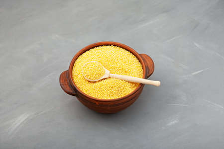 Uncooked Hulled millet in clay pot. Yellow grain, wooden spoon. Gray background, selective focus.の写真素材