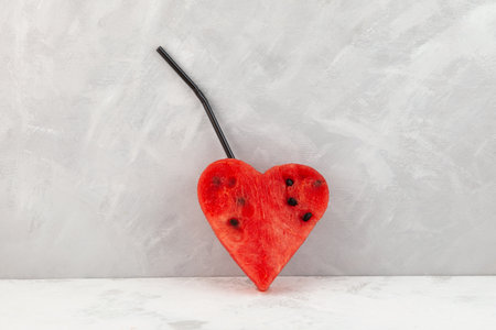 Watermelon pulp in the shape of heart with drinking straw on gray background. Concept of seasonal drinks, refreshing cocktails and smoothies made from fresh watermelon pulp.の写真素材