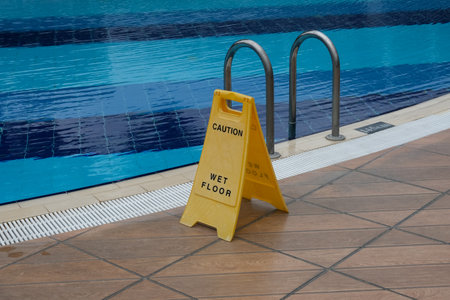 Yellow warning sign with description WET FLOOR near outdoor pool after rain. Slippery ceramic tile floor covering. Concern for the safety of people.の写真素材
