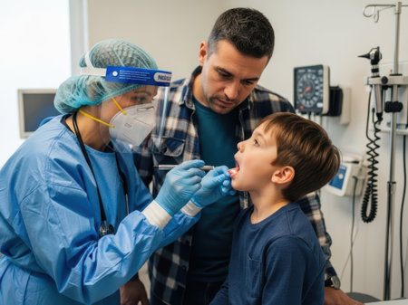 The image depicts a medical professional wearing protective gear, including a face mask and face shield, examining a young boy's throat. The boy is seated and appears to be cooperative. The scene takes place in a clinical setting with medical equipment visible in the background. The image is on a transparent background, making it suitable for various digital applications.の素材