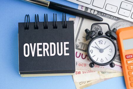 OVERDUE text with keyboard, fountain pen, alarm clock, banknotes currencies and calculator on blue background. Coronavirus Covid19 and Business Conceptの写真素材