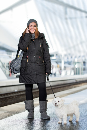 happy young woman having a phone call on her smartphone while waiting with a maltese dog on the leane at the train stationの写真素材