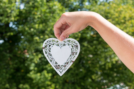 White metal heart hanging down held by hand in a park during summerの写真素材