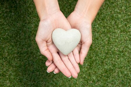 Heart made of stone held in crossed hands above meadow symbolizing care and protectionの写真素材