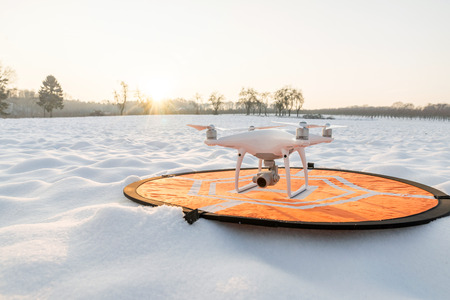Photo of a white professional quadcopter drone camera standing on heliport with white snow in winter in front of forestの写真素材