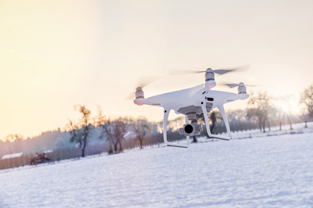 Photo of a flying white professional quadcopter drone camera in the air with white snow in winter in front of forestの写真素材