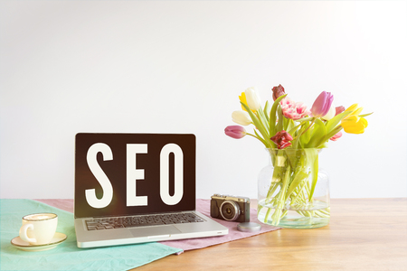 SEO written on laptop screen as abbreviation for search engine optimization for websites on wooden office desk with flowers in front of white backgroundの写真素材