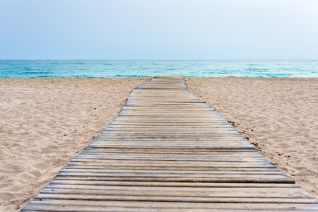 Wooden boardwalk made of planks across the beach to the sea water with sand surrounding as...