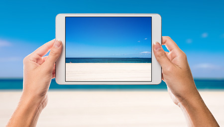 Holiday scene with beach and sea on white tablet screen with hands holding the device up against ocean and beach in the backgroundの写真素材