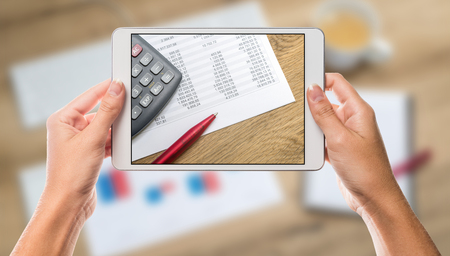 Business calculator and chart paper on white tablet screen with hands holding the device and office chart table in the backgroundの写真素材