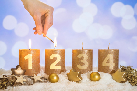 Four numbered gold advent candles with two candles lit by hand with match and christmas decoration lying in snow as templateの写真素材