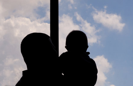 A young man holding a child at the window looks outの写真素材