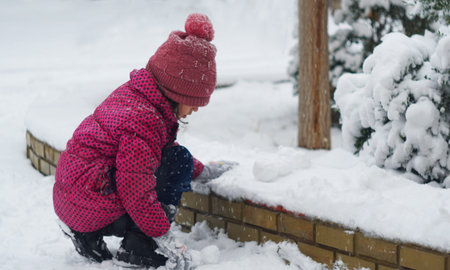 The girl is making a snowball. The girl is wearing a red jacket. She is playing in the snow. It is...