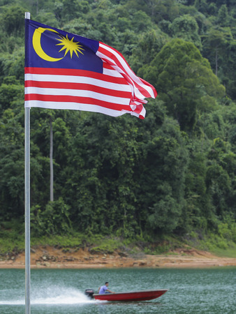 Malaysia Flag, Jalur Gemilang waving with the background of boat at the lake and Malaysian rainforest trees.の写真素材