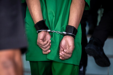 Arrested male criminal in prison uniform and handcuffs is brought by a Police officer to the Law and...