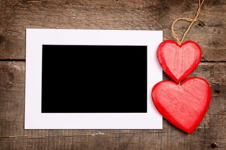 Two red wooden heart shapes with a blank picture frame on rustic woodの写真素材