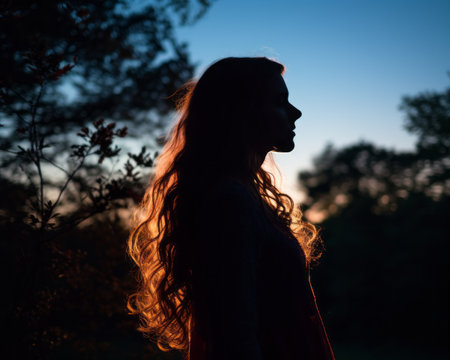 a silhouette of a woman with long hair standing in front of trees at sunsetの素材