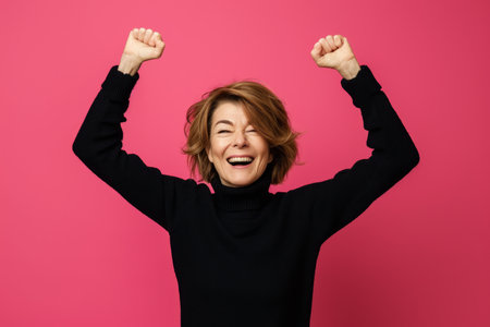 a woman in a black sweater raising her arms in the air on a pink backgroundの素材