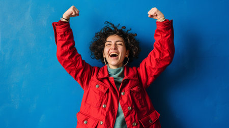a woman in a red jacket is raising her arms up against a blue wallの素材