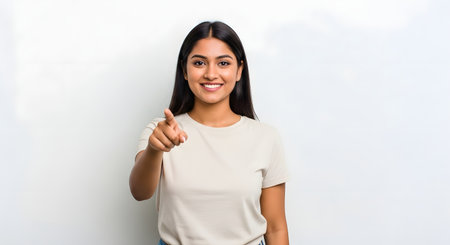 Smiling young Indian woman points directly at viewer. Her confident expression and clean white background create engaging image. She wears simple t-shirt. Brunette looks directly at camera inviting interaction. Direct gaze, modern aesthetic, and cheerful presentation. Professional and reliable.の素材