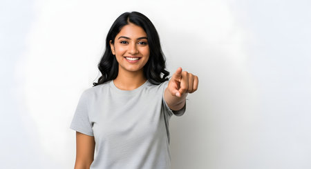 Attractive woman pointing finger at viewer with friendly smile and positive expression. Isolated on white background. Young woman indicating to camera for encouragement and invitation. Confident female points with positive attitude and happiness.の素材