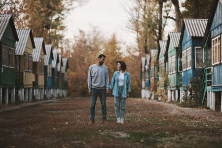 Smiling couple stanting with holding hands between row of wooden cabin and looking to each otherの写真素材
