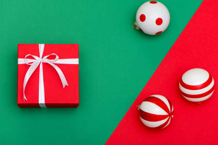 Flat lay of red gift box with white ribbon and Christmas toys on green and red background.