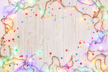 Festive Christmas background with garland lights and colorful stars on white wooden surface.